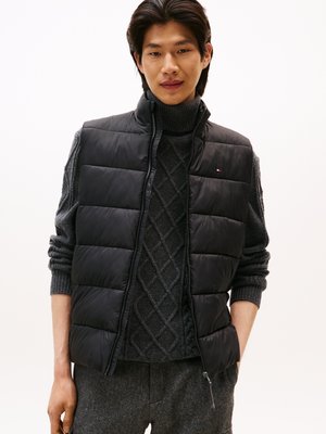 Gilet rembourré noir avec col montant, doté d'une fermeture éclair et d'un logo. Porté par-dessus un pull en maille gris foncé avec un motif en losanges.