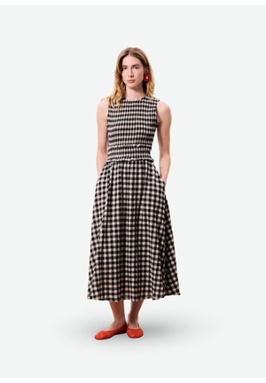 Femme debout, les mains dans les poches, portant une robe sans manches à carreaux noirs et blancs et des chaussures plates rouges avec des boucles d'oreilles fleurs rouges.