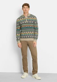 Strickpullover in gemustertem Beige, Grün und Marine mit halbem Reißverschluss am Kragen; kombiniert mit beigen Hosen und hellen Sneakers. Lässige Passform und strukturierter Stoff.