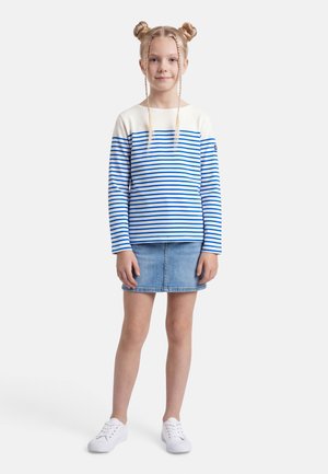 Fille aux cheveux blonds coiffés en deux chignons, portant un t-shirt à manches longues rayé bleu et blanc, une jupe en jean, et des baskets blanches, debout sur un fond blanc.