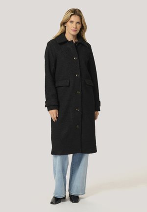 Manteau en laine noir avec une finition texturée, un col, des poches avant et une fermeture à boutons. Associé à un jean large bleu clair et des bottes noires.