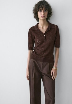 Jong persoon met krullend haar draagt een bruin gebreid shirt met korte mouwen en losse bruine broek, staand tegen een eenvoudige lichte muur.