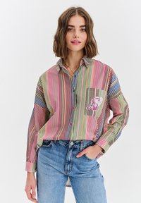 Płaszczowy, w paski, koszula z wielokolorowego materiału ma długie rękawy, kieszeń na piersi z graficznym wzorem i luźny krój. Nosi się ją z niebieskimi dżinsami.