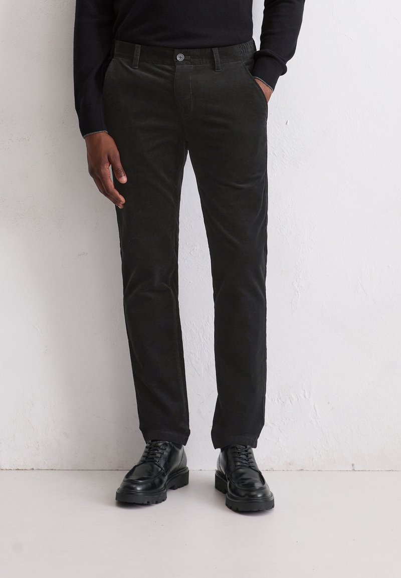 Homme portant un pantalon en velours côtelé noir, des chaussures noires à lacets et un haut noir à manches longues, debout contre un mur blanc.