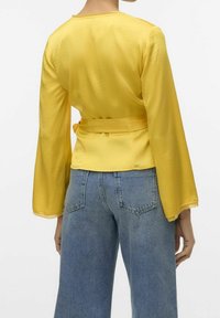 Žlutý dlouhý rukáv top se stahovací šňůrkou v pase, vyrobený z hladkého materiálu s krajkovým lemem na konci rukávů. Kombinováno s modrými džíny.