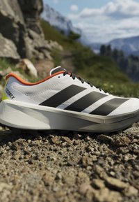Chaussure de randonnée blanche avec des rayures noires et une doublure orange reposant sur un sentier rocheux en milieu montagnard en plein air.