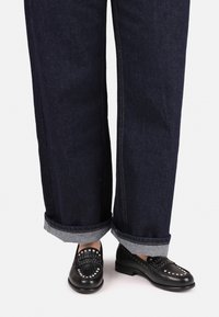 Jeans a gamba larga di colore blu scuro con orli risvoltati, abbinati a mocassini neri con dettagli in metallo argentato e una texture in pelle liscia.
