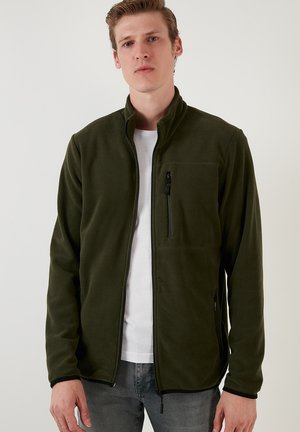 Green fleece jacket with a high collar, zippered pockets, and a smooth texture. Features black zippers and a fitted design.