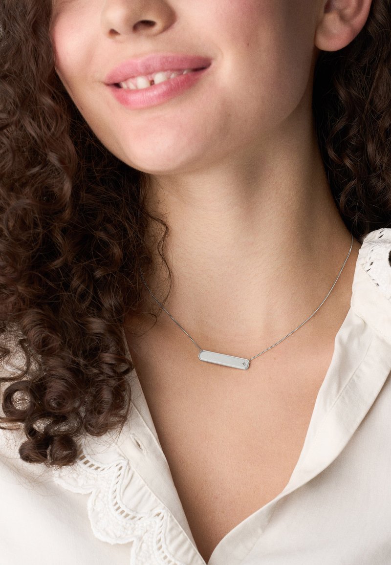 Persona sorridente con capelli ricci che indossa una collana con pendente rettangolare argentato e una blouse bianca con rifiniture in pizzo.