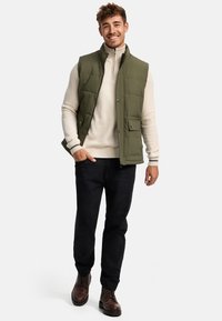 Vesta umplută de culoare verde măslin peste un pulover crem, pantaloni negri și bocanci cârți. Prezintă o închidere cu fermoar și buzunare laterale.