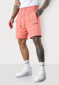 Shorts corail avec taille à cordon, dotés de poches latérales et d'un logo. Associés à des baskets blanches et des chaussettes rayées.