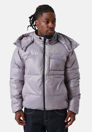 Man met dreadlocks die een lichtgrijze donsjas draagt met een borstzak en ritssluiting, kijkend naar de zijkant tegen een witte achtergrond.