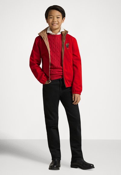 Piros cipzáros dzseki bézs gallérral, alatta piros kötött pulóver, fekete nadrág és fekete cipő; tiszta, szabott megjelenés.