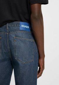 Blue denim jeans with a smooth texture, featuring a visible back pocket and a blue HUGO logo patch at the waistband. Black shirt in background.
