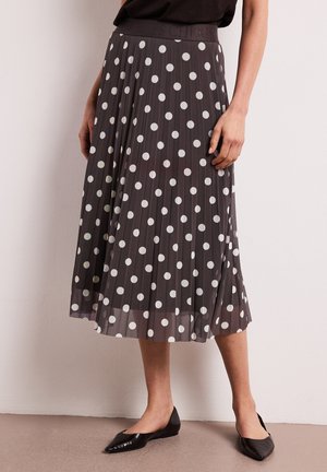 Frau trägt einen schwarzen plissierten Midirock mit weißen Punkten und schwarze spitze Ballerinas, steht an einer weißen Wand.