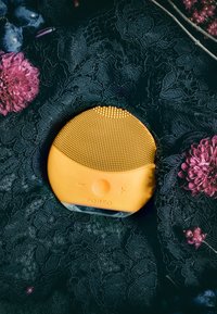 Gul ansiktsrengöringsapparat med texturerad silikonytan, rund form, knappar för drift, placerad på en mörk spetsbakgrund med blommor.