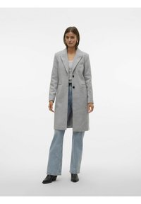 Cappotto grigio in misto lana con design sartoriale, chiusura monopetto, revers e tasche frontali. Indossato sopra un top chiaro e jeans svasati.