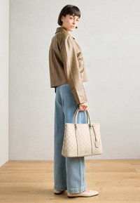 Sac fourre-tout beige avec des accents plus clairs, arborant un motif en diamants et des double poignées. Le modèle porte une veste en cuir marron et un jean bleu.