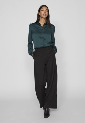 Satin donkerblauwgroene blouse met een kraag, lange mouwen en knoopdetails, gecombineerd met hoge zwarte wijde broek en enkellaarsjes.