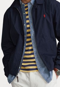 Marineblau leichte Jacke, über einem hellen Denim-Hemd und einem gelb gestreiften Oberteil getragen. Verfügt über einen Reißverschluss, seitliche Taschen und ein kleines Logo.