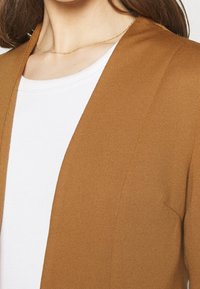 Terrakotowy kardigan z otwartym przodem o prążkowanej fakturze i długich rękawach, noszony na białej koszulce z okrągłym dekoltem. Widoczne subtelne przeszycia.