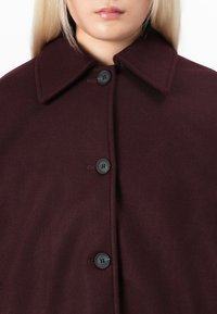 Manteau en laine bordeaux avec un col haut, doté d'une fermeture à bouton et d'une texture douce. Design simple avec des accents de boutons noirs.