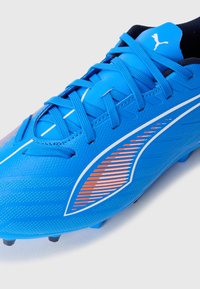Blå Puma fodboldstøvle med blå snørebånd, hvid og orange design på siden, og sorte studs på sålen mod en hvid baggrund.
