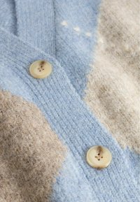 Pehmeä neulottu kangas, jossa on suuria beigejä ja vaaleansinisia laikkuja, ja kaksi marmoroitua beigeä nappia, joissa kummassakin on neljä reikää.