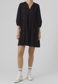 Vestido negro hasta la rodilla con escote en V, mangas abullonadas y diseño en capas. Presenta flecos y detalles texturizados. Se lleva con zapatillas blancas.