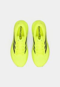 Ζεύγος από φουξ λαμπερά κίτρινα παπούτσια τρεξίματος με μαύρες ρίγες, από πάνω, δεμένα με κορδόνια, με την επιγραφή "SUPERNOVA" ορατή μέσα στα παπούτσια.