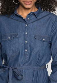Vestido de camisa de denim numa tonalidade azul escura, com fecho de botões à frente, dois bolsos no peito e um cinto de tecido. Textura suave e design de gola clássica.