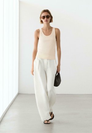 Vrouw loopt binnen, draagt een beige tanktop, witte broek met wijd uitlopende pijpen, zwarte sandalen, zonnebril en houdt een zwarte gestructureerde tas vast.