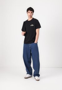 Black cotton t-shirt with a crew neck and chest graphic, paired with loose blue denim jeans. White sneakers complete the look.