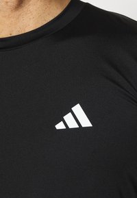 Svart atletisk t-shirt med korta ärmar, tillverkad av ett slätt, elastiskt material. Har en framträdande vit Adidas-logotyp på vänster sida.
