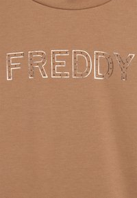 Tessuto marrone con la parola "FREDDY" scritta in lettere metalliche decorate con piccoli puntini neri.