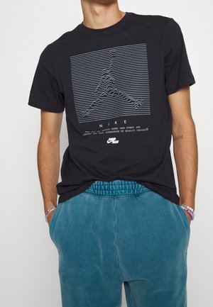 Sort bomulds-T-shirt med et grafisk tryk med bølgede linjer og ordet "NIKE" i hvid, kombineret med grønblå joggerbukser.