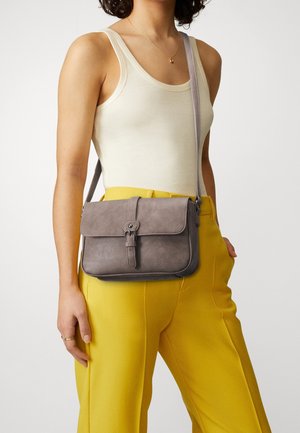 Vrouw met witte mouwloze top en gele broek, draagt een grijze crossbodytas met gespsluiting.