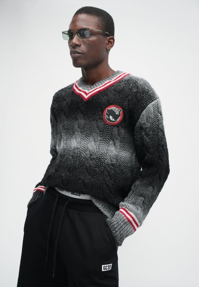 Schwarzer und grauer gestrickter Pullover mit Kabelmuster, V-Ausschnitt, roten und weißen Streifen sowie einem runden Patch. Getragen mit schwarzen Sweatpants.