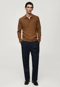 Brown ribbed long-sleeve polo with three buttons, paired with dark blue tailored trousers and black shoes, standing against a neutral backdrop.
