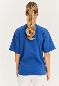 Modrá bavlnená tričko s uvoľneným strihom, krátkymi rukávmi a jednoduchým dizajnom. Zobrazené zozadu, s hladkou textúrou a bez viditeľných log. 