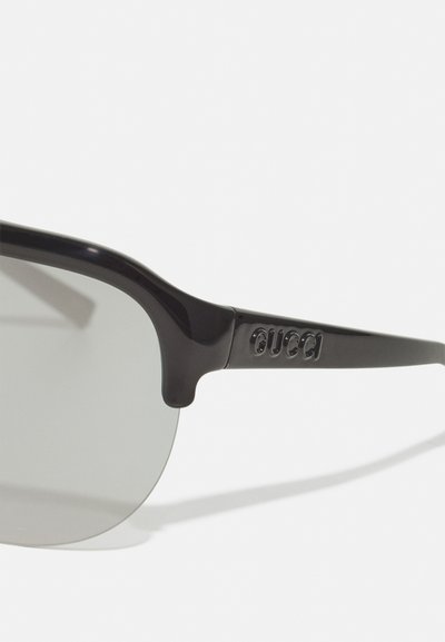 Black plastic sunglasses with gray tinted lenses, featuring a curved frame and embossed logo on the temple. Smooth finish and modern design.
