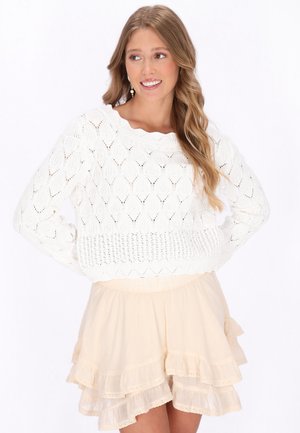 Frau mit langen Haaren, die einen weißen Strickpullover und einen hellbeigen, mehrlagigen Rock trägt, lächelt und schaut zur Seite, während sie die Hände in die Hüften stemmt.