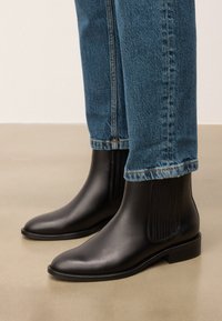 Stivaletti neri in pelle con pannelli laterali texturizzati, punta arrotondata liscia e tacco basso a blocco, abbinati a un paio di jeans in denim azzurro chiaro.