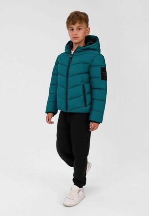 Jeune garçon debout portant une doudoune à capuche bleu sarcelle, un pantalon noir et des baskets blanches sur un fond blanc uni.