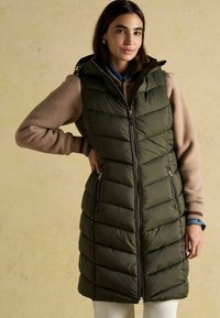 Ciemnozielona kamizelka puchowa z poziomym pikowaniem, kieszeniami na zamek i kapturem. Noszona na jasnobrązowym swetrze, do tego niebieskie dżinsy.