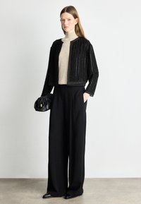 Μαύρο κοντό σακάκι με κάθετη υφή, μπεζ πουλόβερ με ψηλό γιακά και μαύρες παντελόνες με φαρδιά εφαρμογή. Κρατάει μια μαύρη τσάντα με συλλογή, απλά μαύρα παπούτσια.