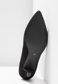 Chaussure à talon haut en daim noir avec une pointe effilée, dotée d'une semelle en caoutchouc noir lisse et d'une étiquette de taille visible "37" sous la chaussure.