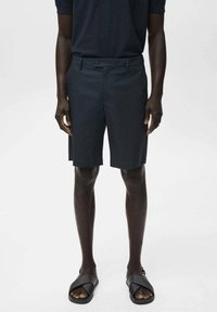 Donkergrijze shorts van een gladde stof, met een sluiting met één knoop en zijzakken. Draagt een paar zwarte slidesandalen.