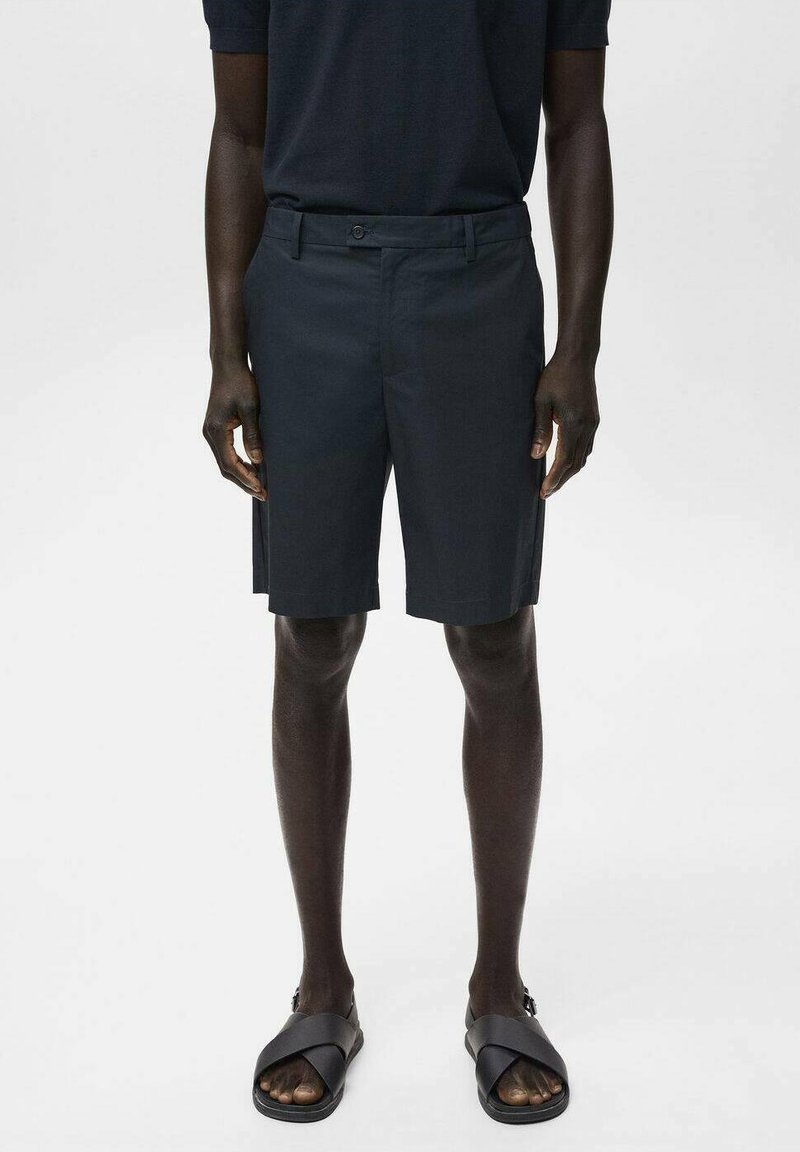 Donkergrijze shorts van een gladde stof, met een sluiting met één knoop en zijzakken. Draagt een paar zwarte slidesandalen.