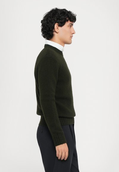 Pull en maille vert foncé avec un col rond et des poignets côtelés, porté sur une chemise blanche à col. Texture lisse, silhouette ajustée.